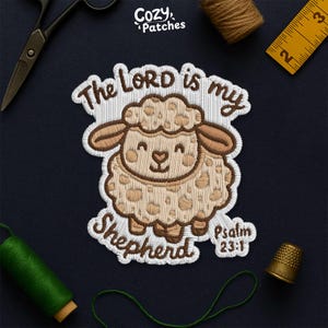 Peut inclure: Écusson brodé représentant un mouton souriant avec le texte "The LORD is my Shepherd" et "Psalm 23:1". Le mouton est beige avec des accents bruns. L'écusson est entouré de coutures blanches. La marque "Cozy Patches" est en haut.