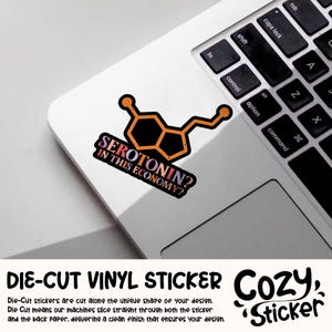K&ouml;nnte beinhalten: Ein gestanzter Vinyl-Aufkleber mit einem schwarzen und orangefarbenen Serotonin-Molek&uuml;l-Design. Der Aufkleber tr&auml;gt den Text "SEROTONIN? IN THIS ECONOMY?" auf einer Laptop-Tastatur. Der Aufkleber ist von Cozy Sticker.