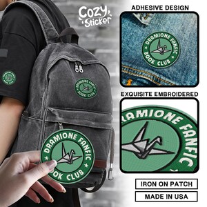 Puede incluir: Un parche bordado verde y blanco con el texto "Dramione Fanfic Book Club" y un dise&ntilde;o de grulla de papel. El parche se muestra en una mochila y una chaqueta vaquera. El parche es para planchar y est&aacute; hecho en EE. UU.