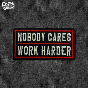 Peut inclure: Un écusson rectangulaire noir avec les mots "NOBODY CARES WORK HARDER" en blanc, soulignés de rouge. L'écusson est sur un tissu texturé gris foncé. Le coin supérieur gauche porte le texte "Cozy Sticker".