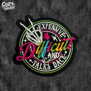 May include: Embroidered patch with the words "Difficult and Talks Back" in a circular design. The patch features a skeleton hand, and the word "Expensive" is also included. The patch has a black background with green, pink, blue, and white accents.