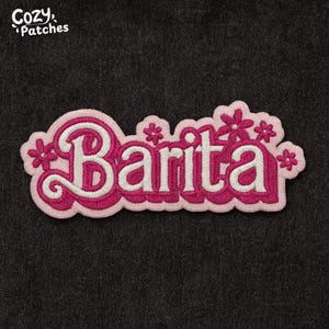 May include: Embroidered patch with the name "Barita" in white script, outlined in pink, and surrounded by a pink border with flower accents. The patch has a textured appearance and the brand "Cozy Patches" is in the top left corner.