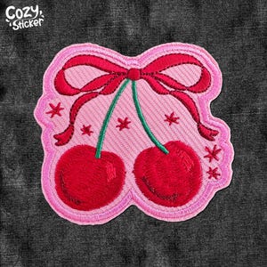 May include: Pink felt patch with an embroidered design of two red cherries with green stems and a red bow. The cherries have white stars around them.