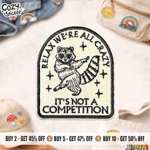 Peut inclure: Un écusson brodé crème avec un raton laveur portant des lunettes de soleil et le texte "RELAX WE'RE ALL CRAZY IT'S NOT A COMPETITION". L'écusson a des coutures noires et une bordure noire. Le logo Cozy Sticker est en haut à gauche.