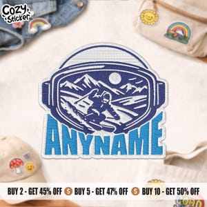 May include: Embroidered patch showing a skier within a goggle-shaped frame, with the text "ANYNAME" in blue. The design includes mountains and a sun. The patch is surrounded by other patches and accessories.