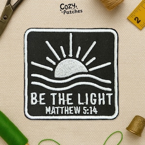 Peut inclure: Écusson brodé noir et blanc avec un soleil et des vagues. Le texte "BE THE LIGHT MATTHEW 5:14" est affiché en dessous. L'écusson est carré avec des coins arrondis.