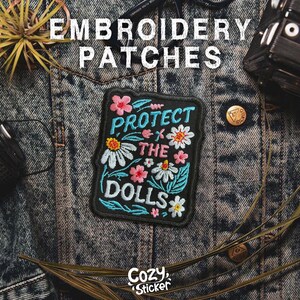 May include: An embroidered patch with the words "PROTECT THE DOLLS" surrounded by colorful flowers. The patch is black with teal, pink, white, and yellow accents. The words "EMBROIDERY PATCHES" are above the patch.