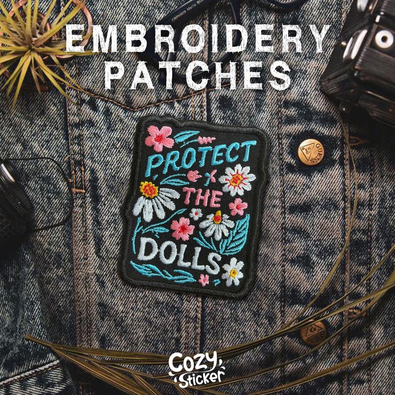 May include: An embroidered patch with the words "PROTECT THE DOLLS" surrounded by colorful flowers. The patch is black with teal, pink, white, and yellow accents. The words "EMBROIDERY PATCHES" are above the patch.