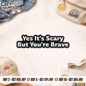 May include: A black and white embroidered patch with the words "Yes It's Scary But You're Brave". The patch is displayed with other accessories, such as a denim jacket and a baseball cap. The brand name "Cozy Sticker" is visible.