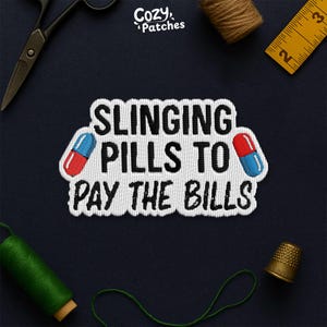 May include: Embroidered patch with the text "SLINGING PILLS TO PAY THE BILLS" in black, with red and blue pill graphics. The patch is white with a black border. The brand name "Cozy Patches" is in the top right corner.