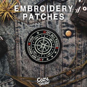 May include: Black and white embroidered patch with a compass rose design and the text "NOT ALL THOSE WHO WANDER ARE LOST." The patch is round and set against a denim jacket. The word "EMBROIDERY PATCHES" is at the top.