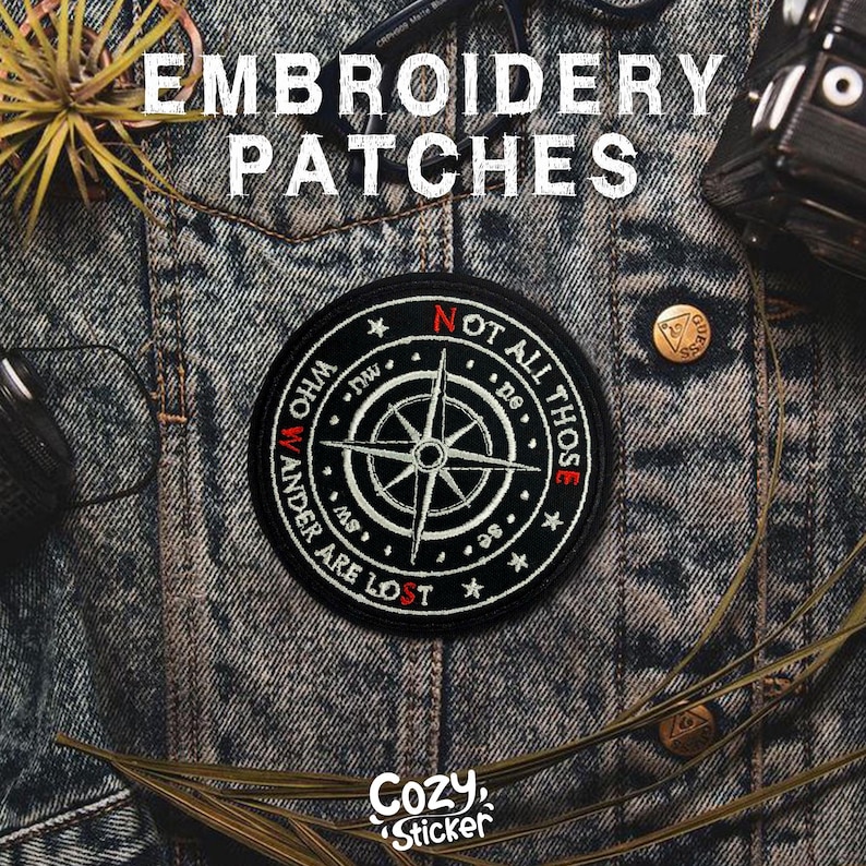 May include: Black and white embroidered patch with a compass rose design and the text "NOT ALL THOSE WHO WANDER ARE LOST." The patch is round and set against a denim jacket. The word "EMBROIDERY PATCHES" is at the top.