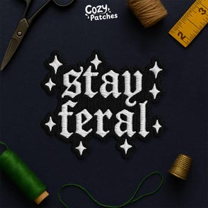May include: Embroidered black patch with the words "stay feral" in white gothic lettering, surrounded by white starburst accents. The patch is on a dark blue background, with sewing tools and thread visible.