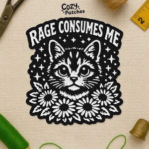 May include: Black and white embroidered patch featuring a cat face with the text "RAGE CONSUMES ME". The cat is surrounded by flowers and stars. The patch is from Cozy Patches.