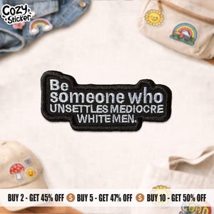 May include: A black patch with white embroidered text that reads "Be someone who unsettles mediocre white men." The patch has a black border and is likely an iron-on or sew-on patch.