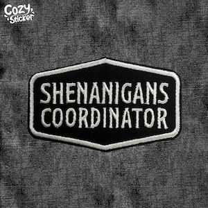 May include: Black and white embroidered patch with the text "SHENANIGANS COORDINATOR" in a bold, sans-serif font. The patch has a black background with a white border and is in a unique hexagonal shape. The patch is on a dark gray fabric.