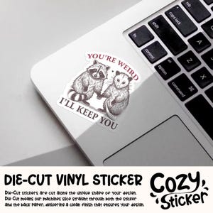 May include: A die-cut vinyl sticker featuring a raccoon and opossum holding hands, with the text "YOU'RE WEIRD I'LL KEEP YOU". The sticker is on a laptop, with the keyboard visible. The sticker is a Cozy Sticker.