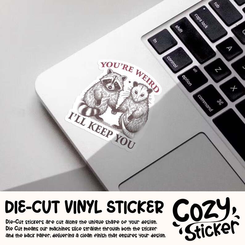 May include: A die-cut vinyl sticker featuring a raccoon and opossum holding hands, with the text "YOU'RE WEIRD I'LL KEEP YOU". The sticker is on a laptop, with the keyboard visible. The sticker is a Cozy Sticker.
