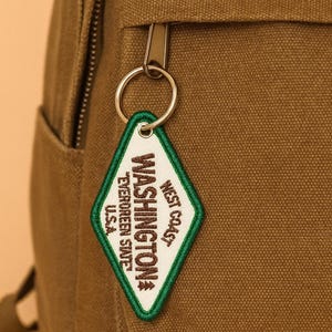 May include: A close-up of a brown backpack with a silver zipper and a diamond-shaped keychain. The keychain has a white background with the words "WASHINGTON" and "WEST COAST" in green and black text.