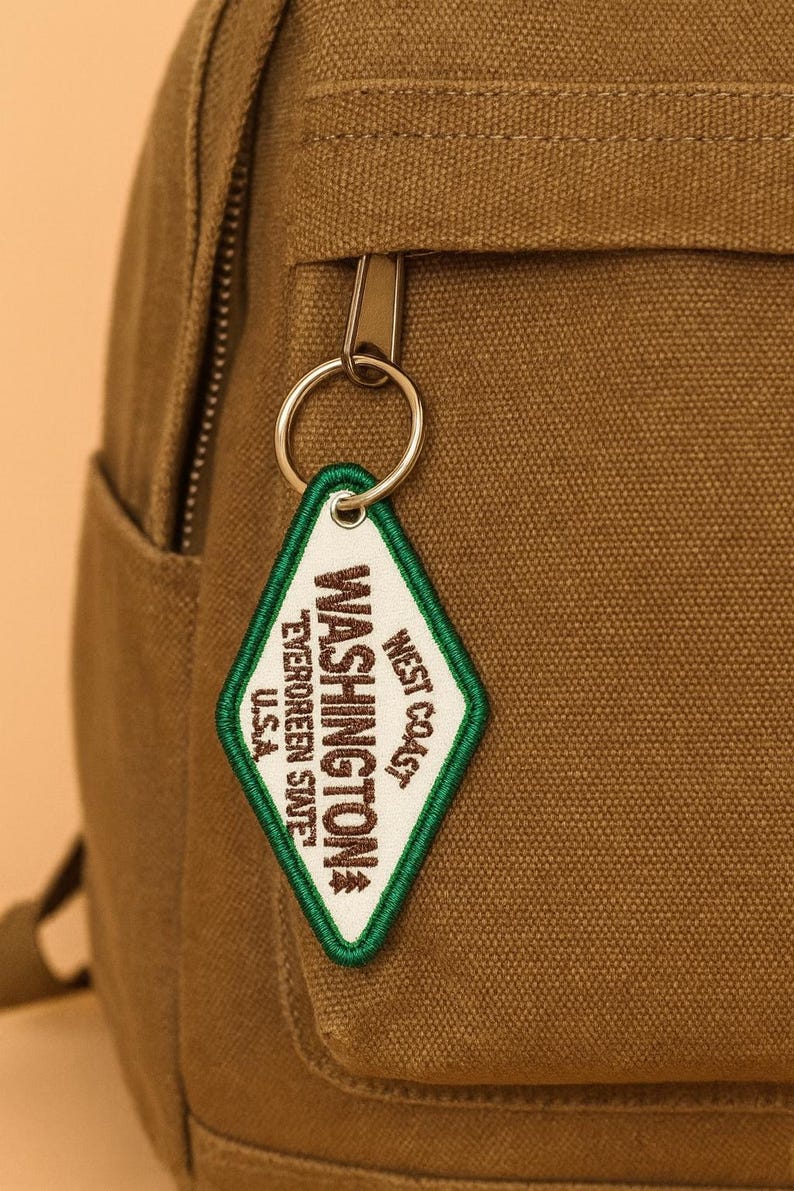May include: A close-up of a brown backpack with a silver zipper and a diamond-shaped keychain. The keychain has a white background with the words "WASHINGTON" and "WEST COAST" in green and black text.