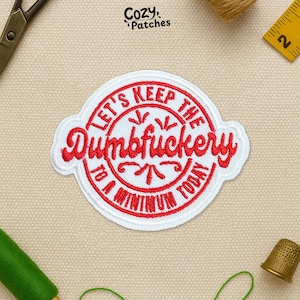 May include: A white and red embroidered patch with the text "LET'S KEEP THE Dumbfuckery TO A MINIMUM TODAY". The patch has a circular design with a decorative border and is surrounded by sewing supplies.