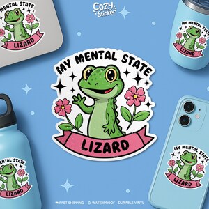 May include: A sticker featuring a cartoon lizard with the text "My Mental State" and "Lizard". The green lizard is waving and surrounded by pink flowers. The sticker is displayed on a laptop, phone, and water bottle, all against a blue background.