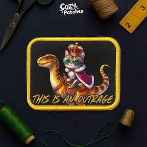 May include: Embroidered patch featuring a regal cat wearing a crown and cape, riding a dinosaur. The patch has a gold border and the text "THIS IS AN OUTRAGE" in yellow. The patch is from Cozy Patches.