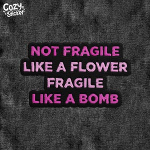May include: A black embroidered patch with the text "NOT FRAGILE LIKE A FLOWER FRAGILE LIKE A BOMB" in pink thread. The patch has a black border and is set against a dark gray, textured background. The "Cozy Sticker" logo is in the upper left corner.