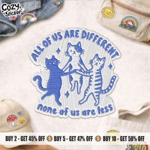 Peut inclure: Un écusson brodé représentant trois chats se tenant par la patte en cercle. Le texte bleu indique ALL OF US ARE DIFFERENT en haut et none of us are less en bas. Les chats sont représentés en fil bleu sur un fond blanc.