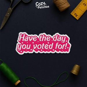 May include: Embroidered patch with the text "Have the day you voted for!" in white on a pink background with a white border. The patch is surrounded by sewing tools, including scissors, thread, and a thimble, on a dark blue surface.