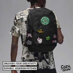 Puede incluir: Mochila negra adornada con varios parches, incluido un c&iacute;rculo verde que dice "Dramione Fanfic Book Club". Otros parches incluyen una pelota de baloncesto, el n&uacute;mero 25 y la palabra "Champions". Tambi&eacute;n se ve el texto "Unleash Your Creativity" y "Durable, Adhesion Patches".