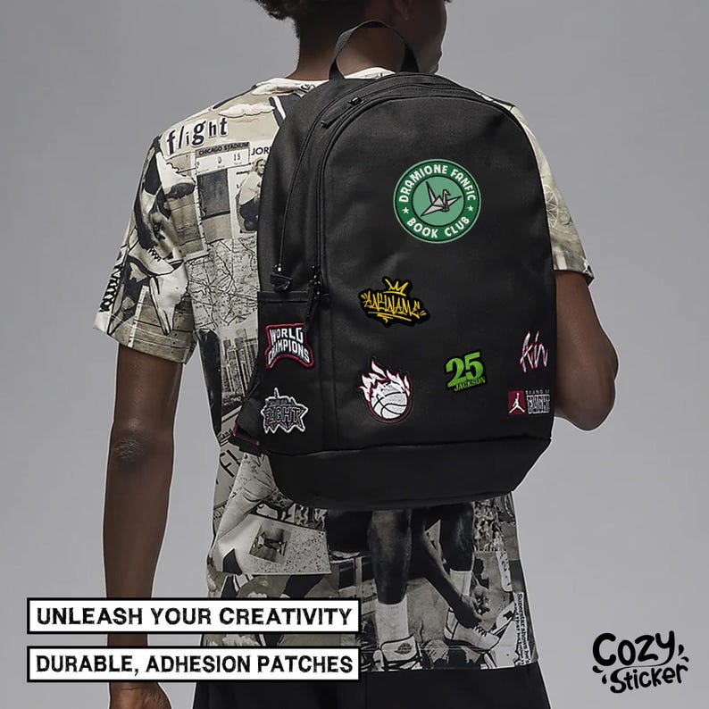 Puede incluir: Mochila negra adornada con varios parches, incluido un c&iacute;rculo verde que dice "Dramione Fanfic Book Club". Otros parches incluyen una pelota de baloncesto, el n&uacute;mero 25 y la palabra "Champions". Tambi&eacute;n se ve el texto "Unleash Your Creativity" y "Durable, Adhesion Patches".