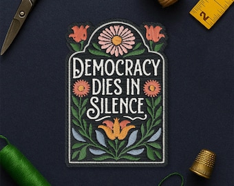 May include: Embroidered patch with the text "DEMOCRACY DIES IN SILENCE" in white script. The patch features floral designs in orange, pink, and green. The patch is surrounded by sewing tools, including scissors, a thimble, and measuring tape.