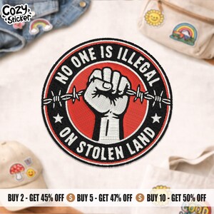 May include: A circular patch featuring the words "NO ONE IS ILLEGAL ON STOLEN LAND" around a clenched fist grasping barbed wire. The patch has a red centre, black border, and white text and fist.