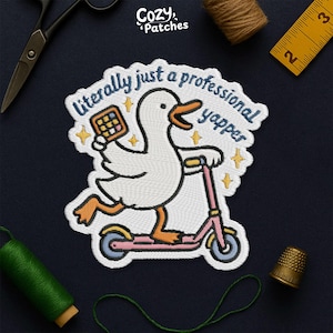 May include: Embroidered patch with a white duck on a pink scooter, holding a phone. Text reads "literally just a professional yapper". The patch is surrounded by yellow stars. The patch is a fun, quirky design.