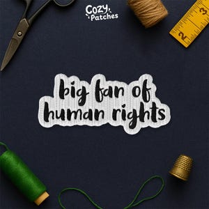 May include: A white embroidered patch with the text "big fan of human rights" in black cursive lettering. The patch is surrounded by sewing tools, including scissors, a measuring tape, and spools of thread, all on a dark blue surface.