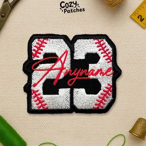 May include: Embroidered baseball-themed patch featuring the number "23" in white, with a black outline and red stitching details. The word "Anyname" is scripted in red across the centre. The patch is from "Cozy Patches".