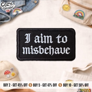 May include: A black rectangular patch embroidered with the white phrase "I aim to misbehave" in a gothic font. The patch is displayed with other accessories, including a denim jacket and a mushroom-themed baseball cap.