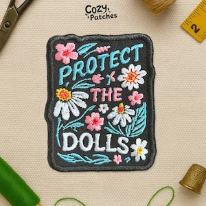 May include: A black embroidered patch with the words "PROTECT THE DOLLS" in white, light blue, and pink. The patch features floral designs with pink and white flowers and green leaves. The patch is rectangular with rounded corners.