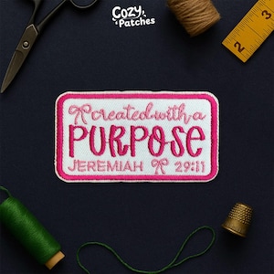 May include: A rectangular patch with a white background and a pink border. The patch features the words "Created with a PURPOSE" in pink script, with "JEREMIAH 29:11" below. The patch is surrounded by sewing tools and supplies.