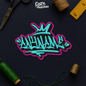 May include: Embroidered patch with the word "APHNAM" in a graffiti style, featuring a black base with teal and pink accents. The patch has a crown design at the top and is surrounded by sewing tools, including scissors, thread, and a thimble.