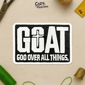 May include: A rectangular black and white patch with the word "GOAT" in large white letters, a cross in the "O", and the text "GOD OVER ALL THINGS." The patch is surrounded by sewing supplies.