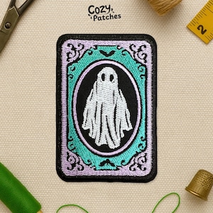 May include: Rectangular patch with a black border, featuring a white ghost inside a black oval. The oval is surrounded by a teal frame with ornate purple detailing. The text "Cozy Patches" is in the upper right corner.