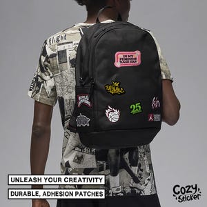May include: Black backpack adorned with colorful patches, including text that reads "IN MY FEMININE RAGE ERA" and "WORLD CHAMPIONS." The backpack has a side pocket and is worn over the shoulder. The image also includes the text "UNLEASH YOUR CREATIVITY" and "DURABLE, ADHESION PATCHES."