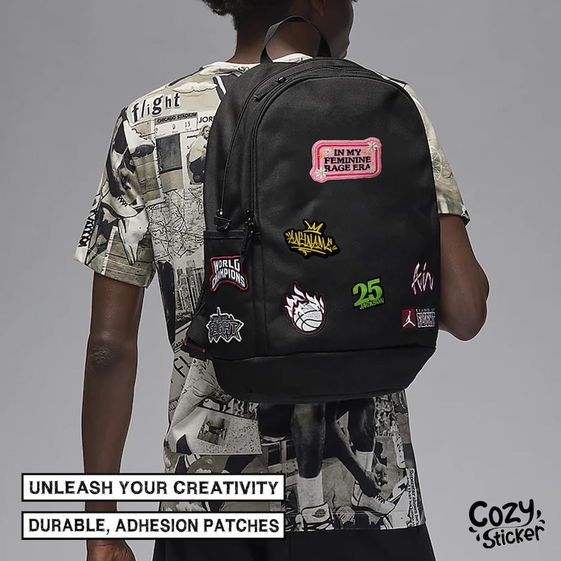 May include: Black backpack adorned with colorful patches, including text that reads "IN MY FEMININE RAGE ERA" and "WORLD CHAMPIONS." The backpack has a side pocket and is worn over the shoulder. The image also includes the text "UNLEASH YOUR CREATIVITY" and "DURABLE, ADHESION PATCHES."