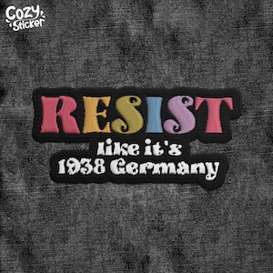 May include: A black patch with the word "RESIST" in colorful embroidered letters. The text "like it's 1938 Germany" is also embroidered in white.