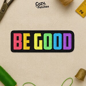 May include: Embroidered patch with the words "BE GOOD" in vibrant rainbow colors on a black background. The patch has a rectangular shape with rounded corners. The brand name "Cozy Patches" is in the top right corner.