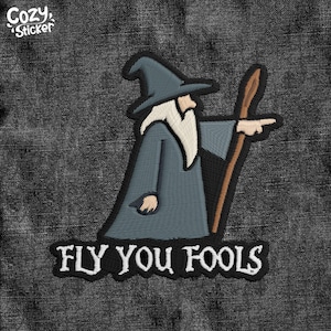 May include: Embroidered patch featuring a wizard in a blue robe and hat, with a white beard, pointing with a staff. The text "FLY YOU FOOLS" is below the wizard. The patch has a black border and the text "Cozy Sticker" in the upper left corner.