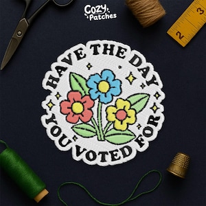 May include: Embroidered patch with the text "Have the day you voted for" surrounding a bouquet of flowers. The flowers are red, blue, and yellow with green leaves. The patch is white with a black border.