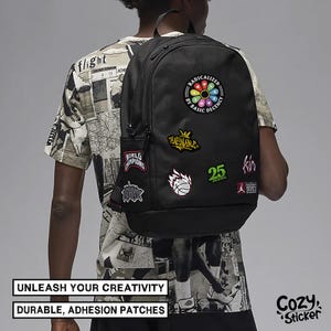 May include: Black backpack adorned with colorful patches, including text that reads "RADICALIZED BY BASIC DECENCY" and "25". The backpack also features patches with basketball and crown designs. The text "UNLEASH YOUR CREATIVITY" and "DURABLE, ADHESION PATCHES" are also visible.